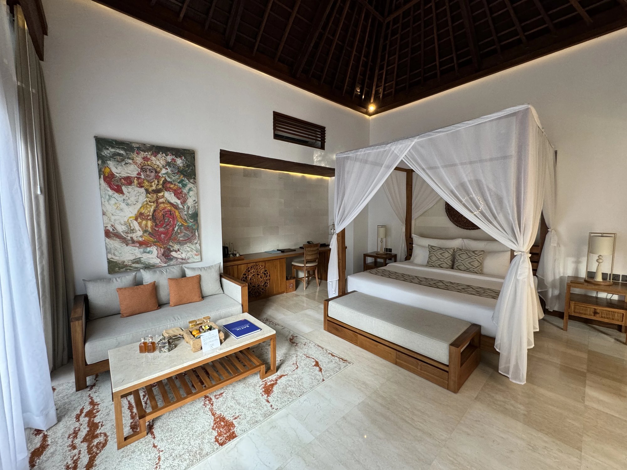 A villa bedroom with a four-poster bed under white netting, a sofa and a large Balinese painting