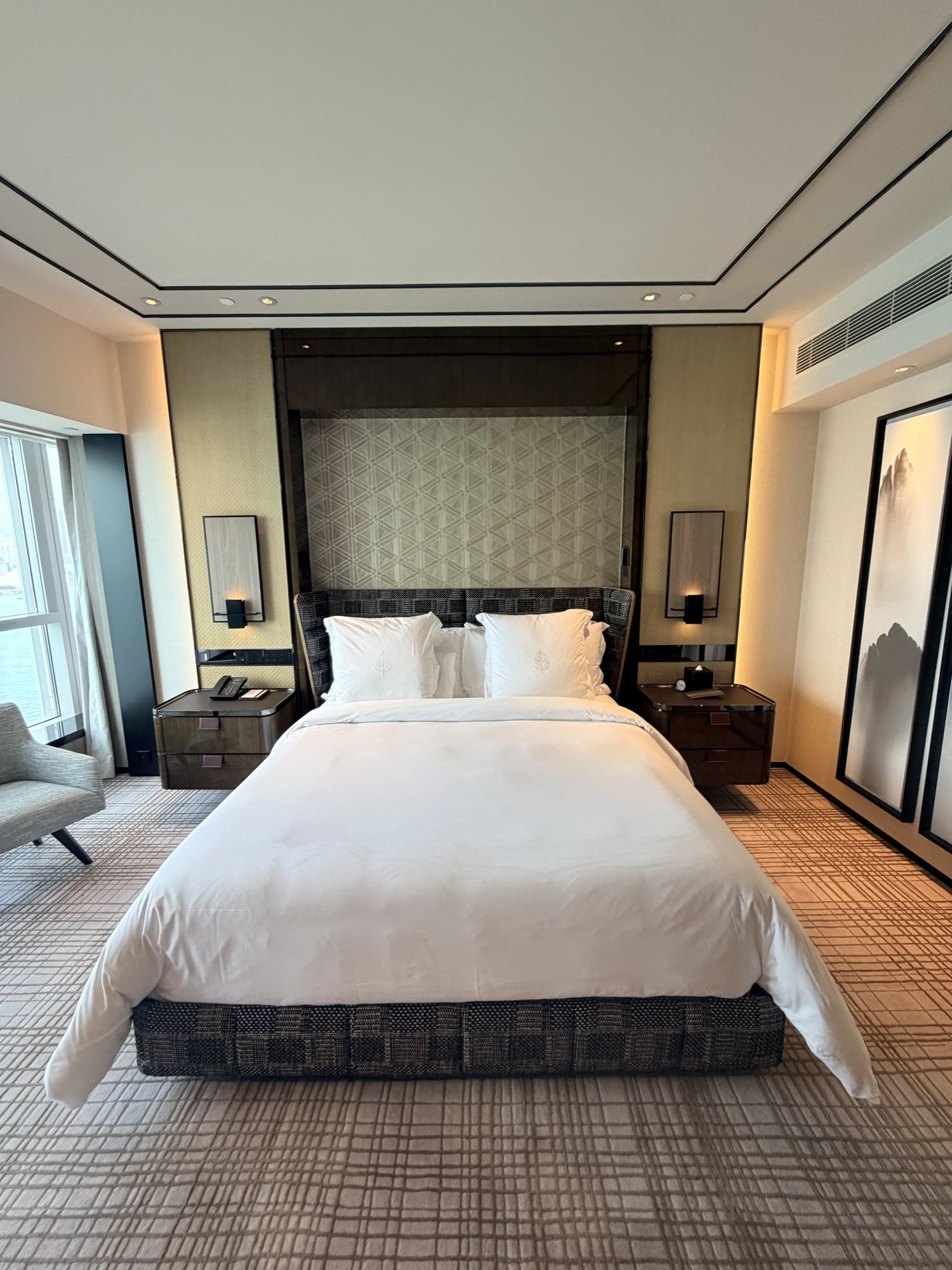 A bed with white linen and a padded woven headboard against a geometric panel, wall lamps on both sides, a grey armchair by the window on the left and an ink landscape painting on the right