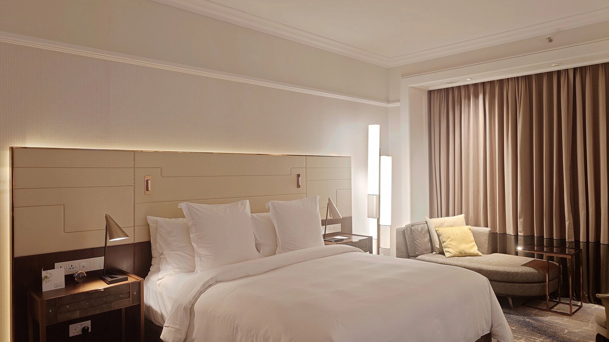 A Deluxe Room with a king bed, reading lamps and a chaise by the curtained window