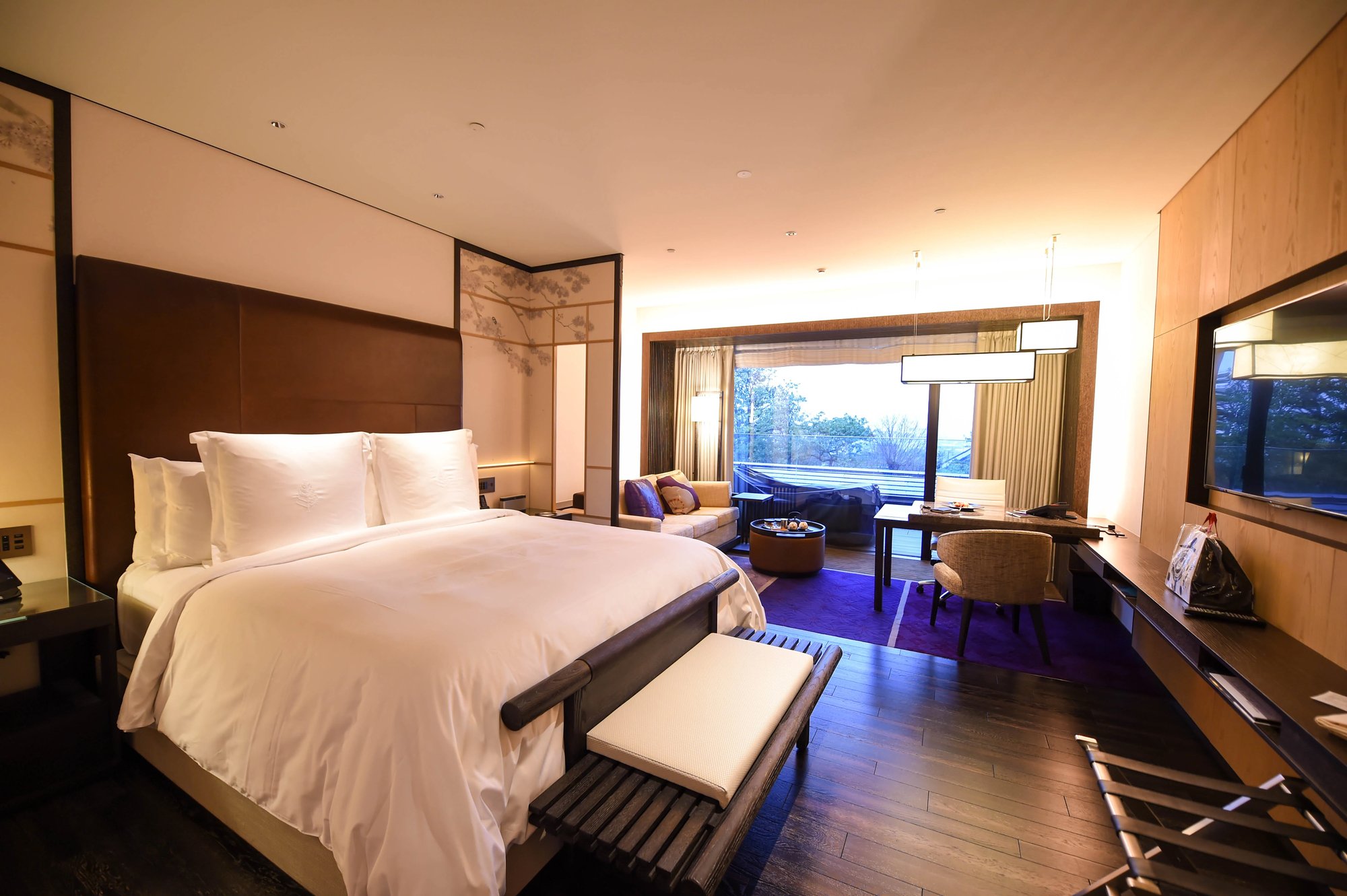 A wide, warmly lit bedroom with a large white bed and a bench at its foot, a sofa with purple cushions on a purple carpet, a desk, and glass doors open to a balcony over treetops