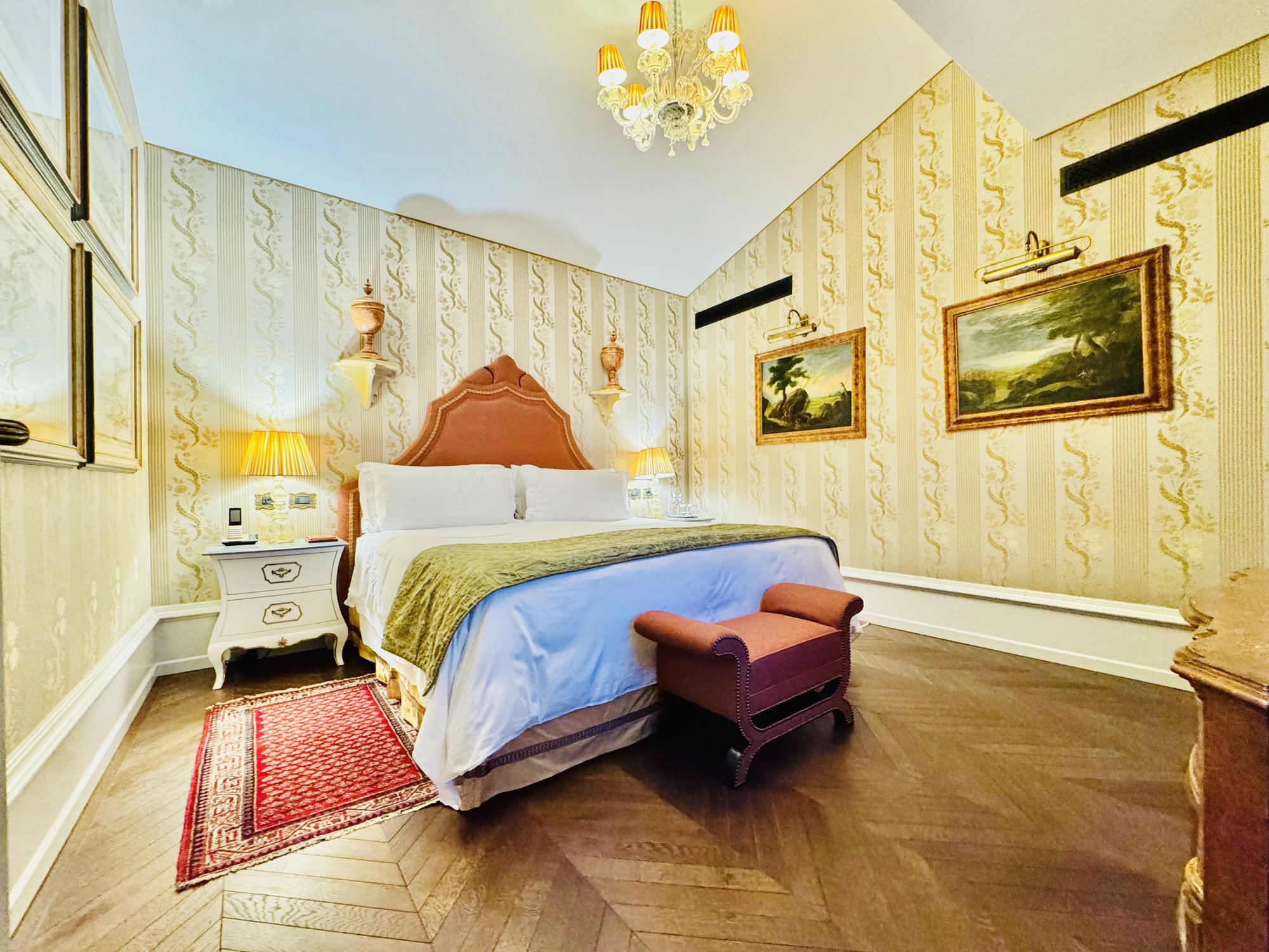 A wide view of the suite bedroom with a king bed, a rust bench at its foot, a chandelier and a sloped ceiling, in bright light