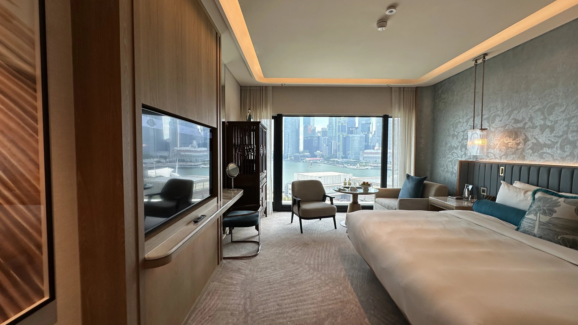 A long hotel room with a TV wall on the left, a king bed against a blue batik-pattern wall on the right, and an armchair and round table at a window over Marina Bay and the skyline