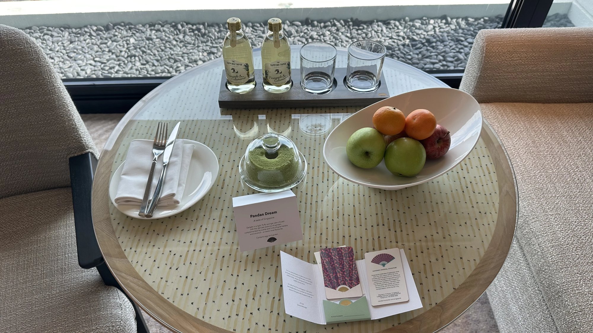 A pandan cake, a fruit bowl and two bottles of sparkling juice on a round table by the window