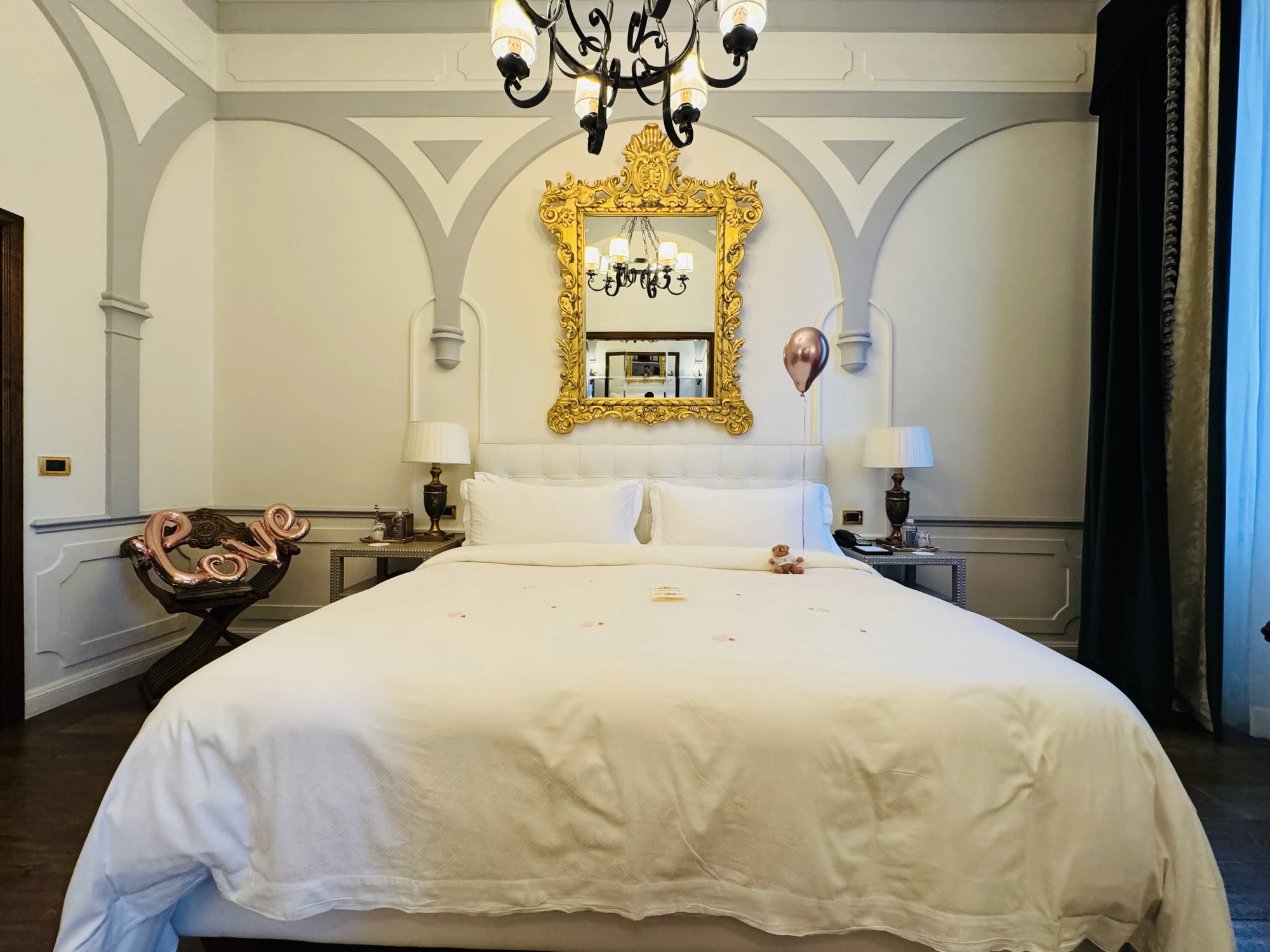 Wide view of the Junior Suite bedroom with a king bed under a gilt mirror, two lamps and a dark green curtain at the tall window.