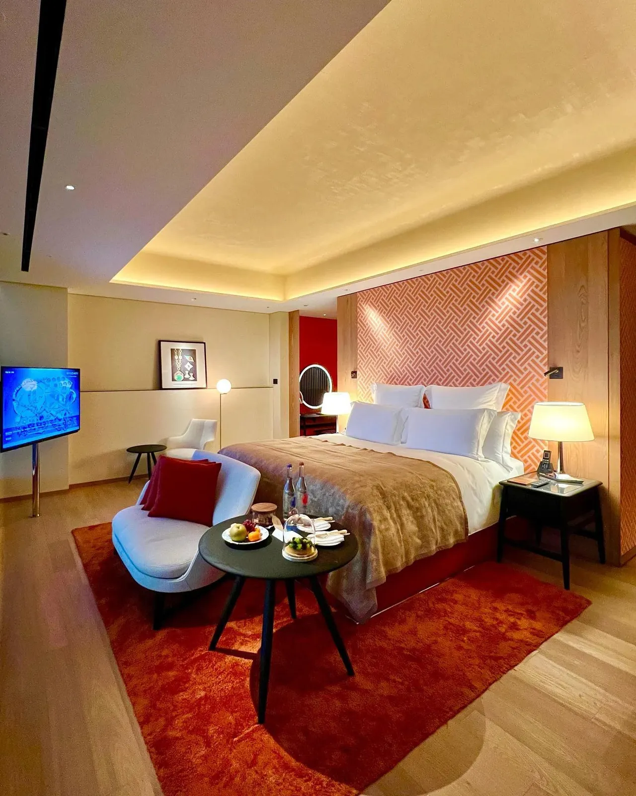 The full room: a wide bed under a glowing cove ceiling, a red rug, a grey chaise and the city through the window
