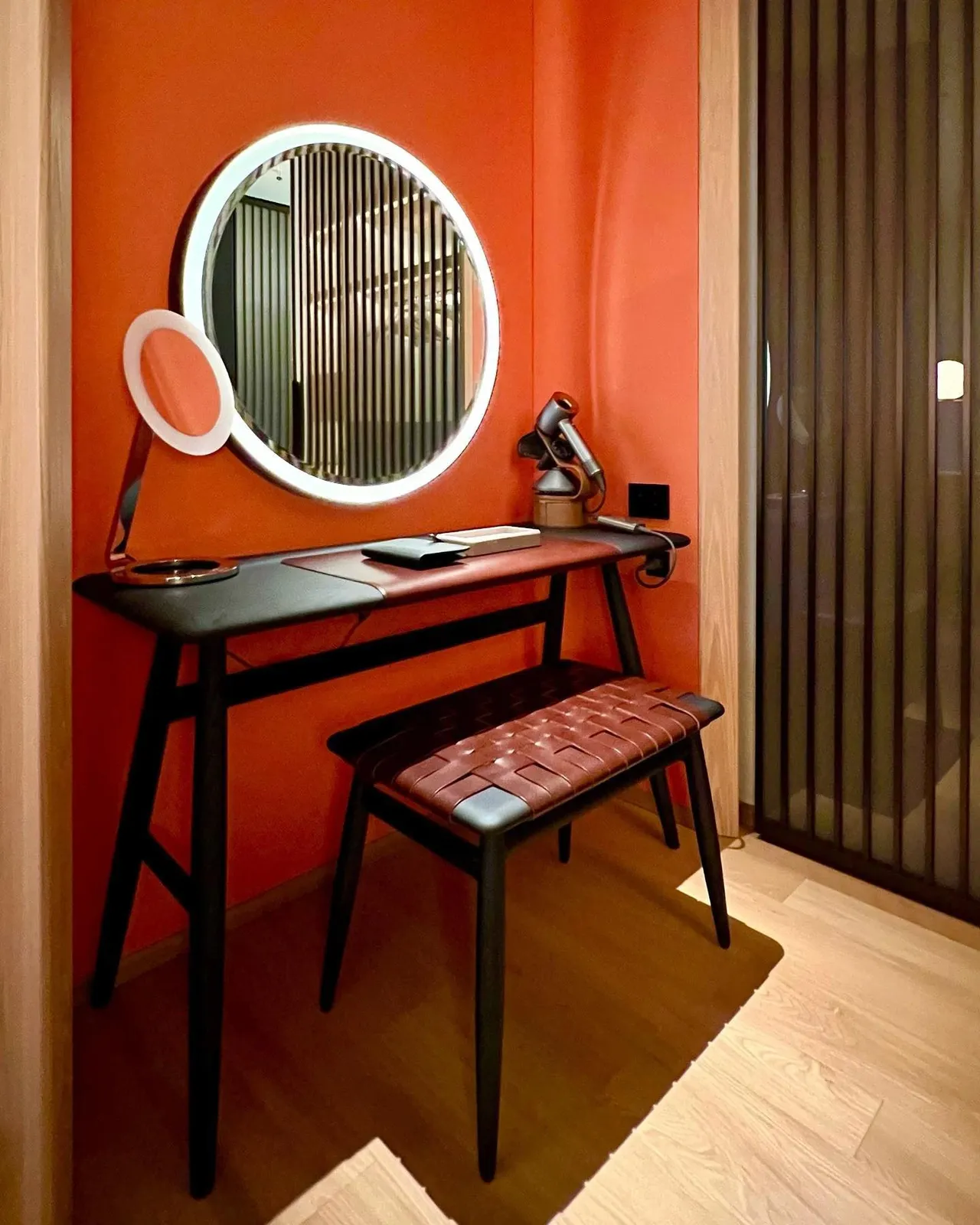A round lit mirror and leather stool at a dressing table against an orange wall
