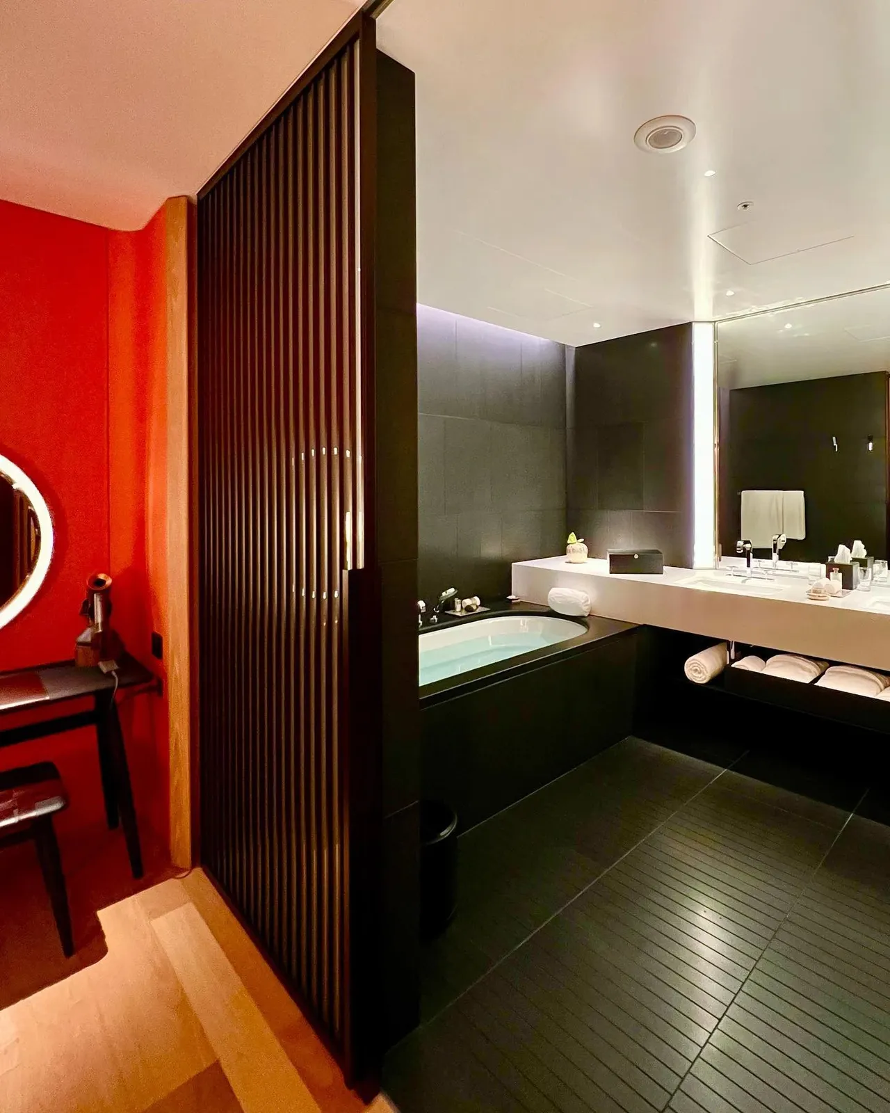 The black stone bathroom seen through the slatted wood screen, with the tub and long vanity