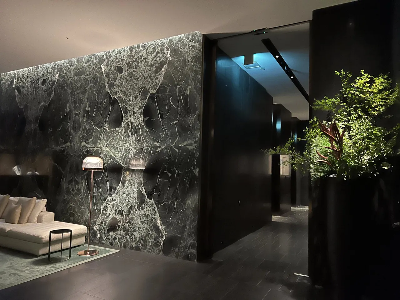 A veined marble feature wall and pale sofa beside the lobby entrance, with a leafy branch in a dark vase