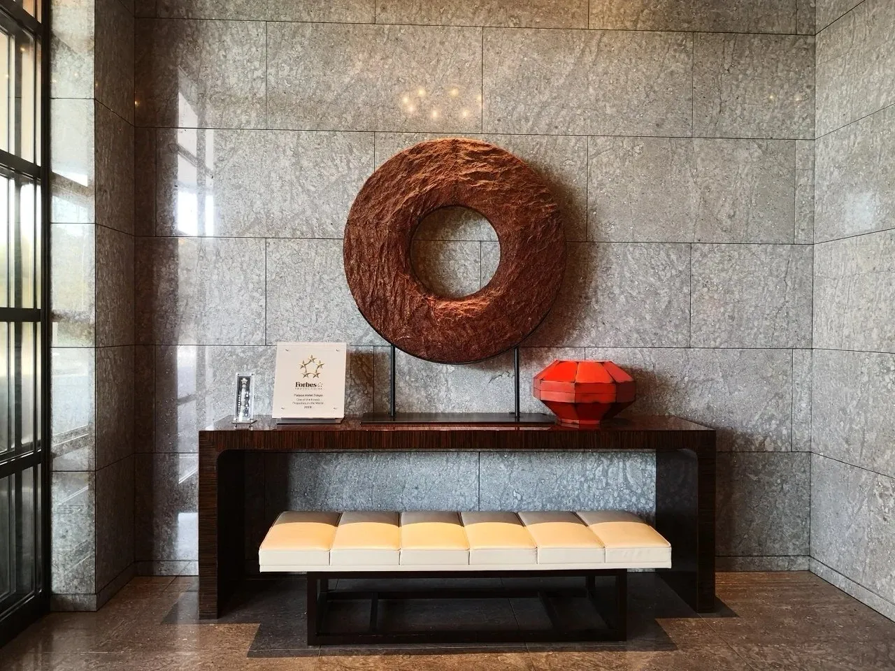 A carved wreath above a dark console and cream bench in a stone-walled lobby corridor