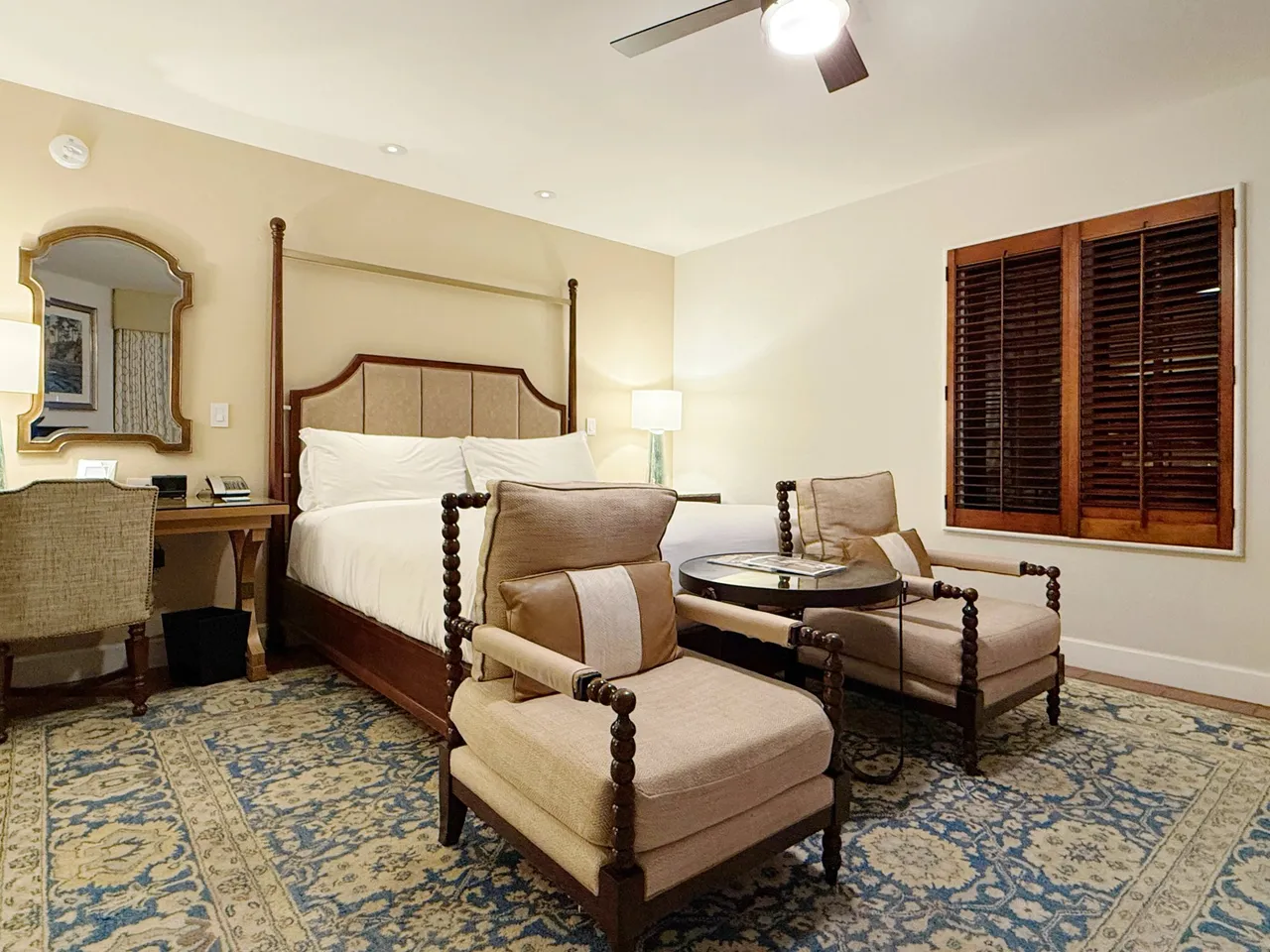 Our Club-level room with a four-poster king bed, two spindle-legged armchairs, a desk with a gilt mirror and wooden shutters, in warm lamplight